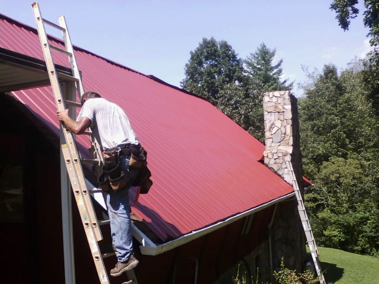 Roofer on a ladder beside a red metal roof and stone chimney — Soffit & Fascia Repair by Roofing Brothers in Cuba